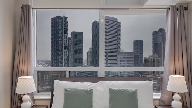 A hotel room with a large window offering a cityscape view, a bed with white and green pillows, and two bedside lamps.