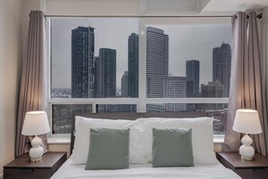 Hotelkamer met groot raam en uitzicht op de stad, een bed met witte en groene kussens en twee nachtlampjes.