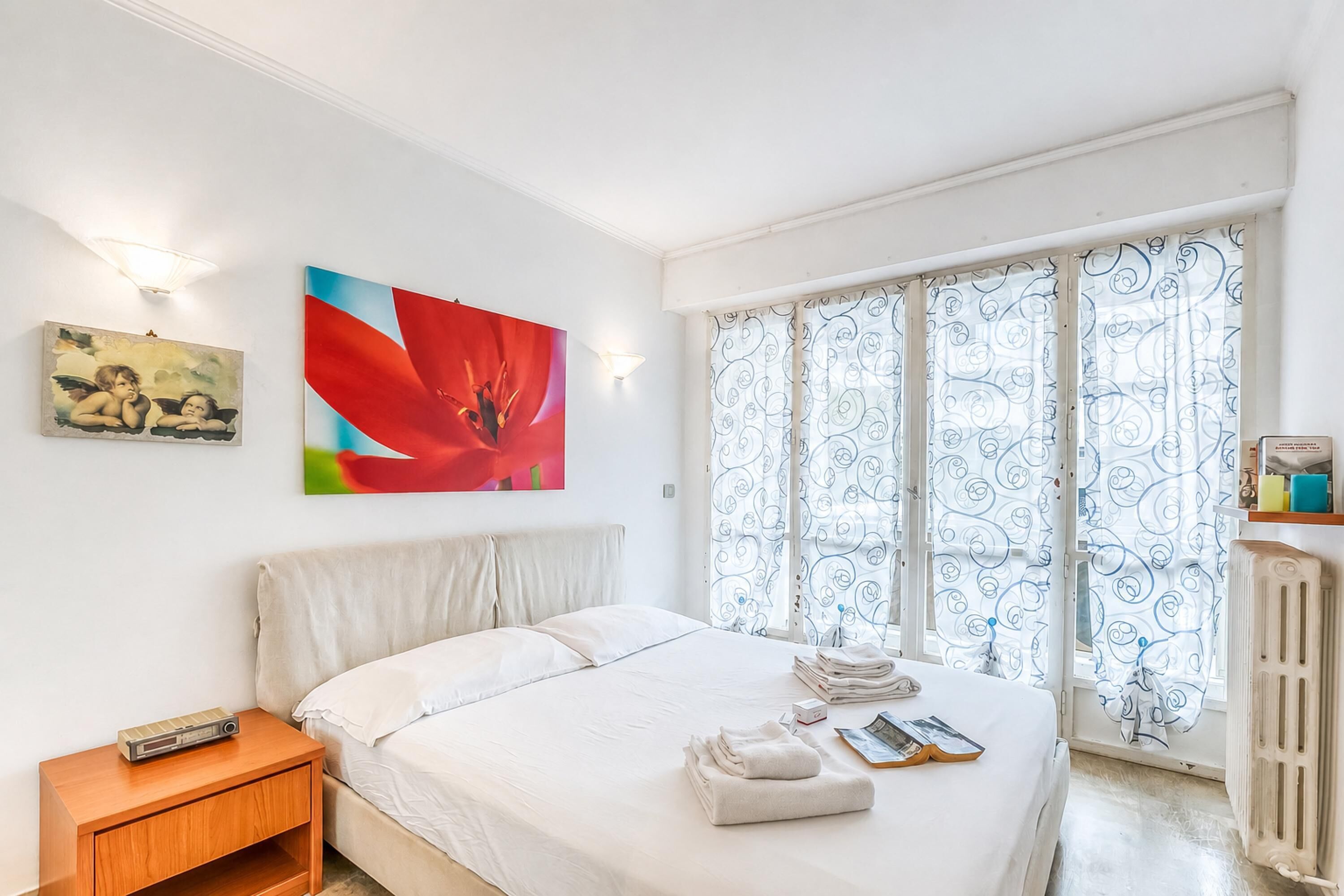 A bedroom with a bed, a nightstand, a painting, a window with curtains, and a radiator.