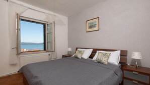 Een slaapkamer met een groot bed, een nachtkastje met een lamp, een raam met uitzicht op zee, en een ingelijste foto aan de muur.