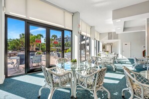 Een hotelkamer met een grote glazen deur die leidt naar een buitenruimte, ingericht met witte rieten stoelen en tafels, en met uitzicht op palmbomen en gebouwen.