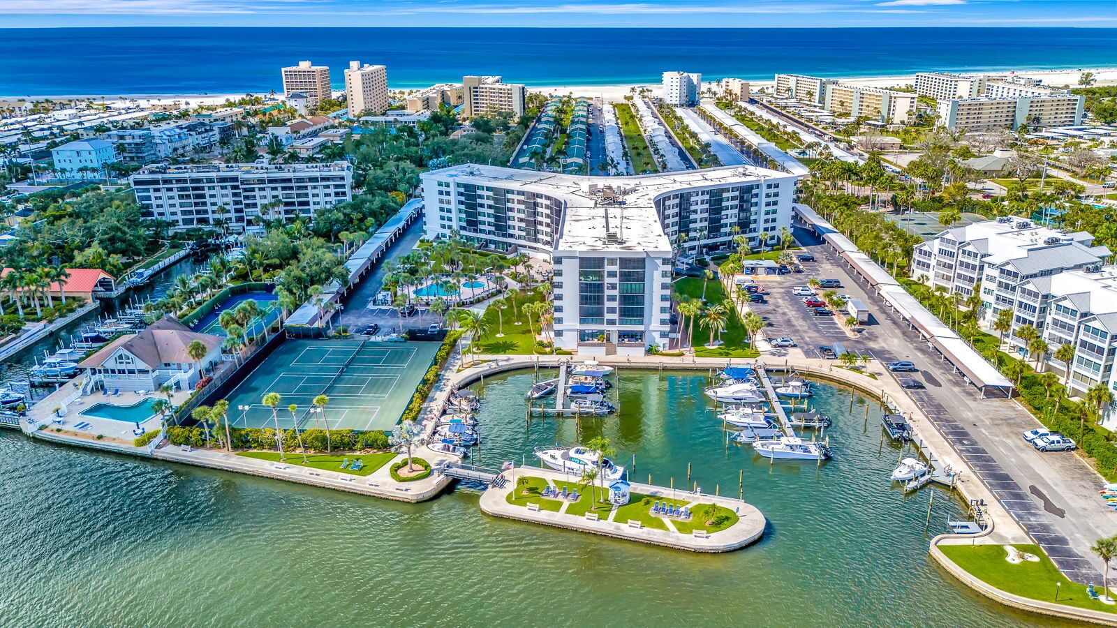 Vue aérienne d’une marina où sont amarrés des bateaux, d’un court de tennis et d’un hôtel à la façade incurvée.
