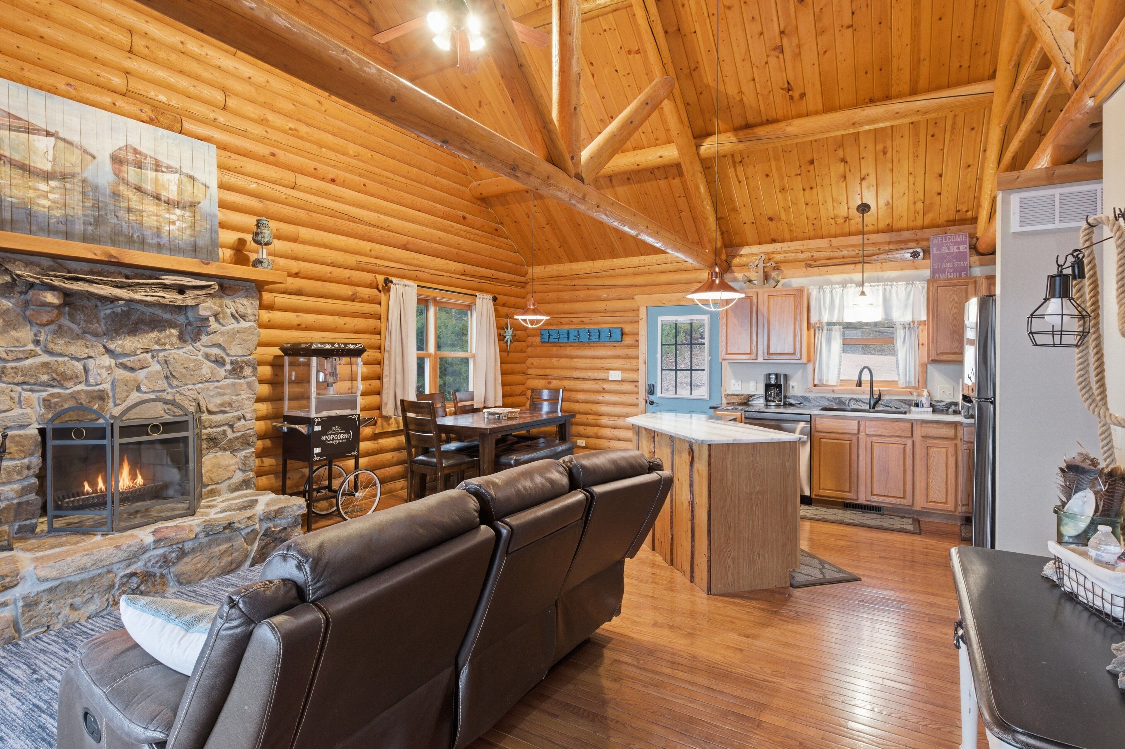 A log cabin interior with a stone fireplace, leather sofa, wooden dining table, and a kitchen area.
