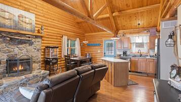 A log cabin interior with a stone fireplace, leather sofa, wooden dining table, and a kitchen area.