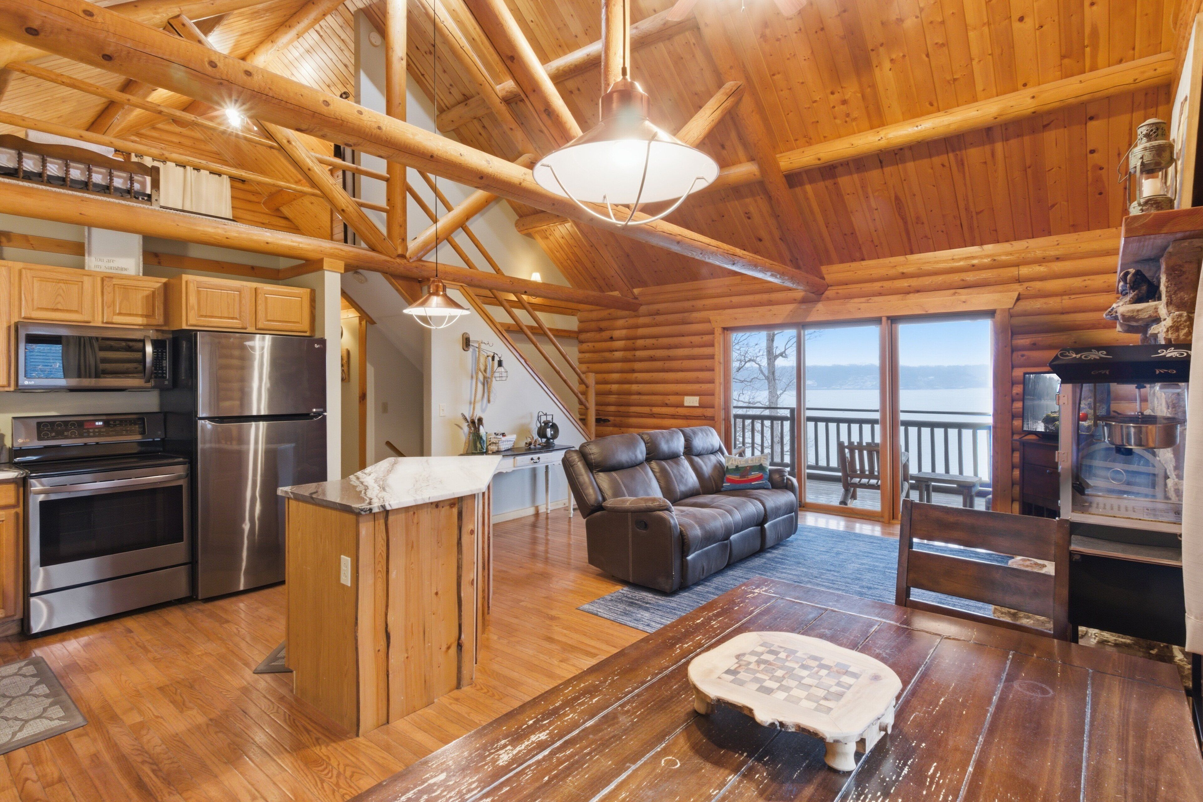 Interior de una cabaña de madera con cocina, comedor y zona de estar con vistas al exterior.