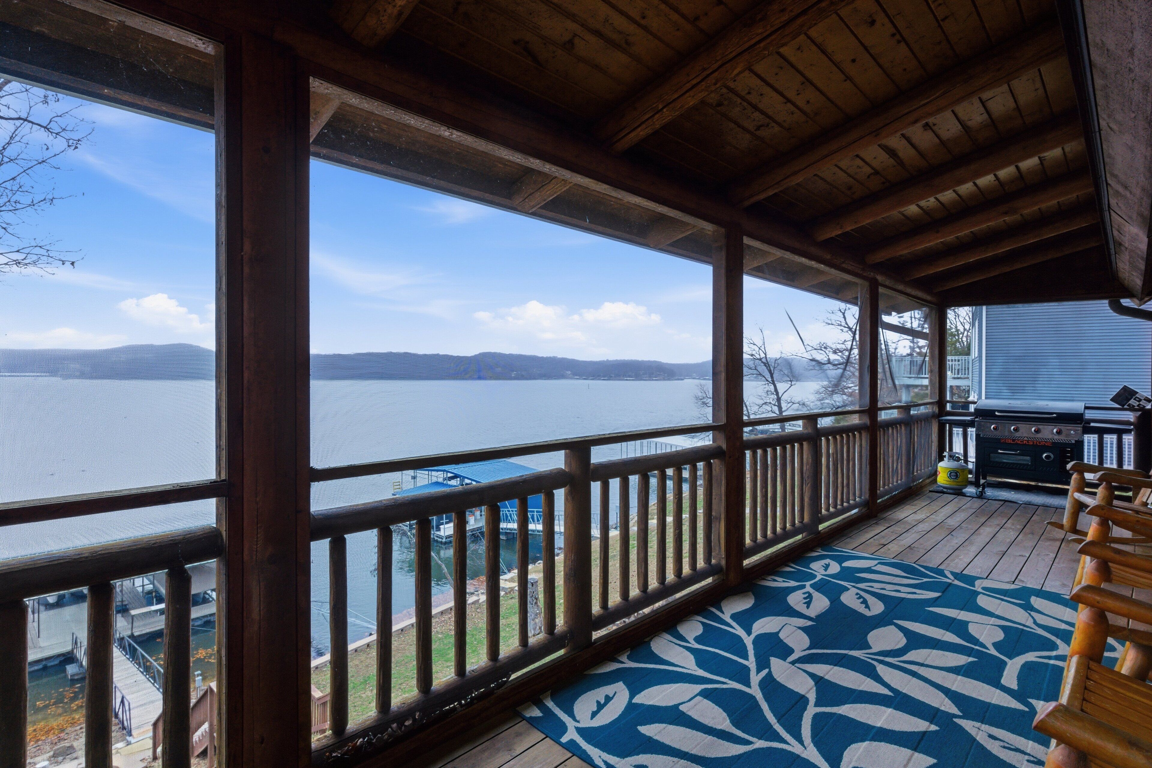 Eine überdachte Terrasse mit Blick auf einen See und Berge, Holzgeländer und Grillplatz.
