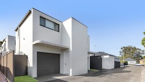 Un edificio residenziale moderno su due piani con un garage, un piccolo giardino anteriore con erba sintetica e una casa vicina.