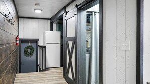 Un pasillo con suelo de madera, una puerta blanca adornada con un adorno navideño, un refrigerador blanco y una reja de metal negro.