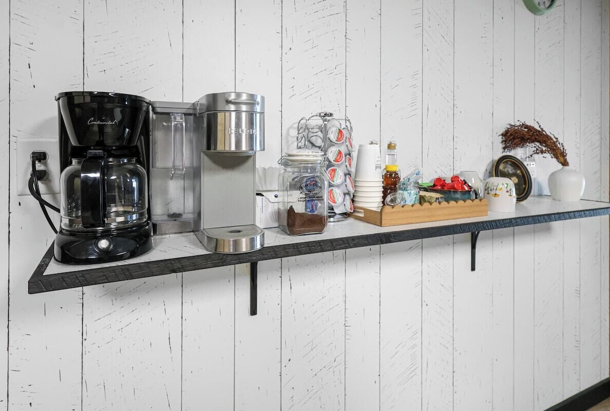 A kitchen counter with a coffee maker, a kettle, a jar of sugar, a honey dispenser, a box of tea bags, and a decorative vase.