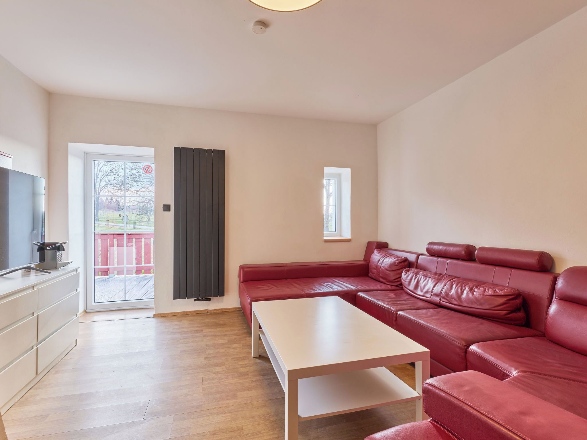 A modern living room with a red leather sofa, a white coffee table, a TV, and a sliding glass door leading to an outdoor deck.