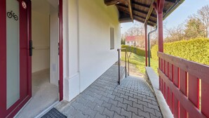 A covered outdoor area with a red railing, a bicycle symbol on a door, and a view of a house in the background. - Růžová 4 by Interhome (Ruzova)