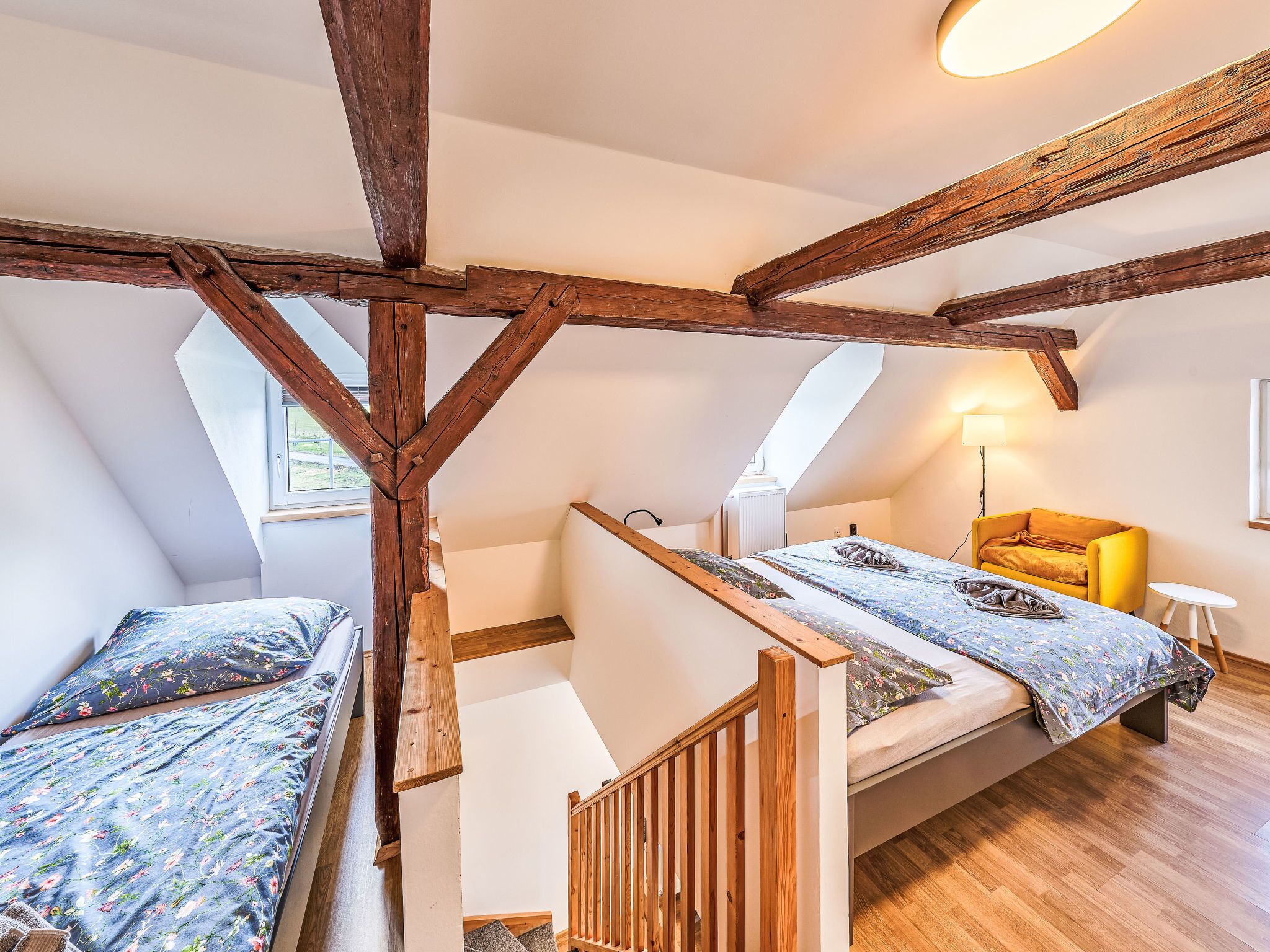A loft bedroom with exposed wooden beams, a bed with floral bedding, a yellow armchair, and a small table.