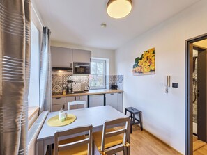 A compact kitchen with a microwave, stove, and sink. There is a dining table with chairs and a small stool. A painting of yellow roses hangs on the wall.