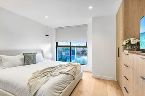 A modern bedroom with a large bed, a view of a swimming pool, and a wooden cabinet with a flat-screen TV. - Luxe on Sandringham San Souci Sanctuary (Sans Souci)