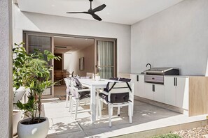 A modern outdoor dining area with a white table and chairs, a ceiling fan, and a kitchen with a grill. - Luxe on Sandringham San Souci Sanctuary (Sans Souci)