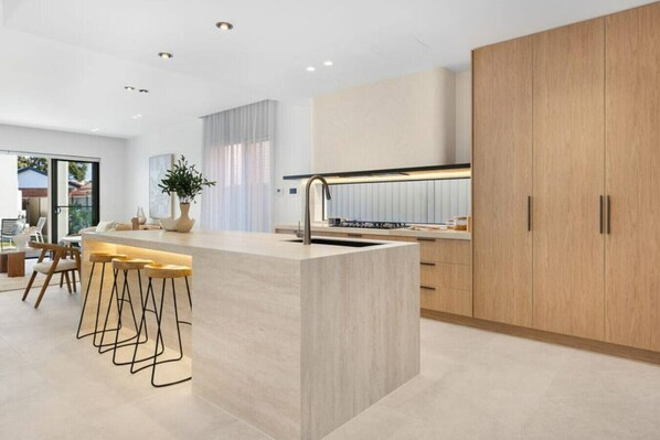 A modern kitchen with a central island, bar stools, and wooden cabinets. - Luxe on Sandringham San Souci Sanctuary (Sans Souci)
