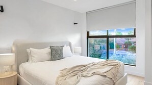 A bedroom with a large bed, a bedside table with a lamp, and a window with a view of a garden and buildings. - Luxe on Sandringham San Souci Sanctuary (Sans Souci)