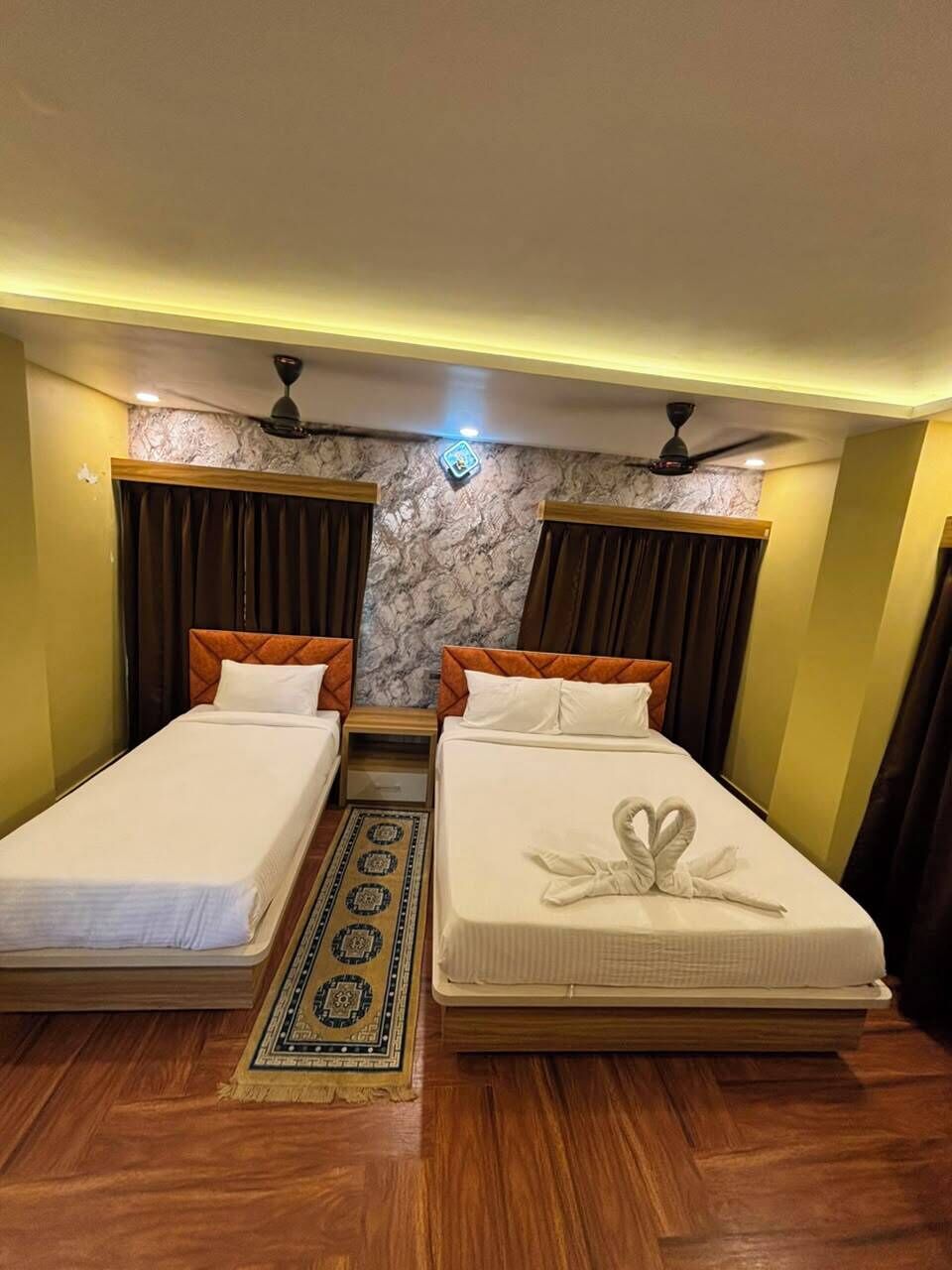 Une chambre d’hôtel avec deux lits simples, un plancher en bois, un mur décoré d’un motif marbré et un ventilateur de plafond.