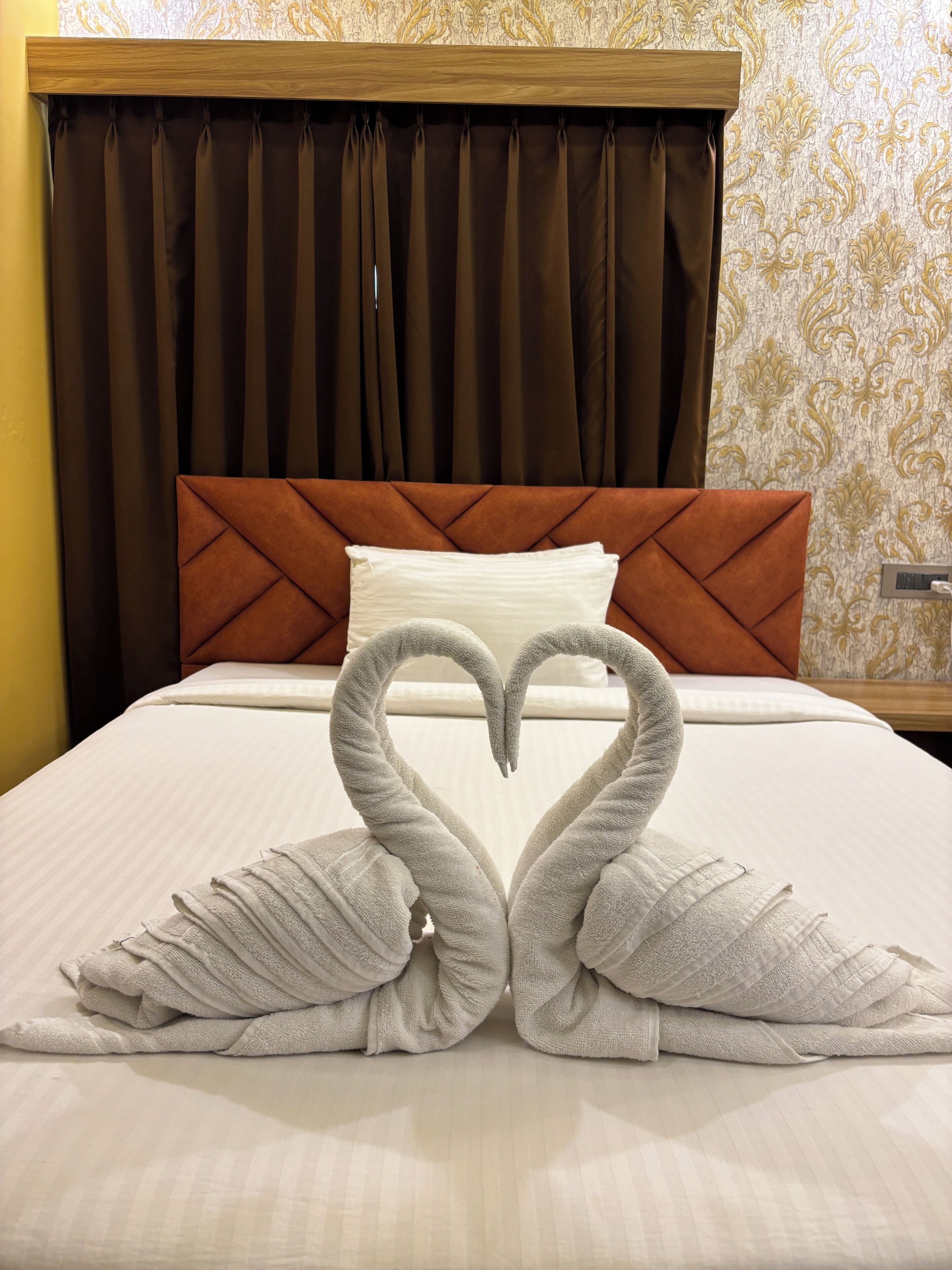 A bed with a heart-shaped towel arrangement, a brown headboard, and a patterned wallpaper.