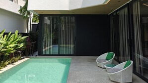 Un área de piscina moderna al aire libre con balaustrada de vidrio, dos sillones de mimbre con cojines verdes y un cielo azul despejado.