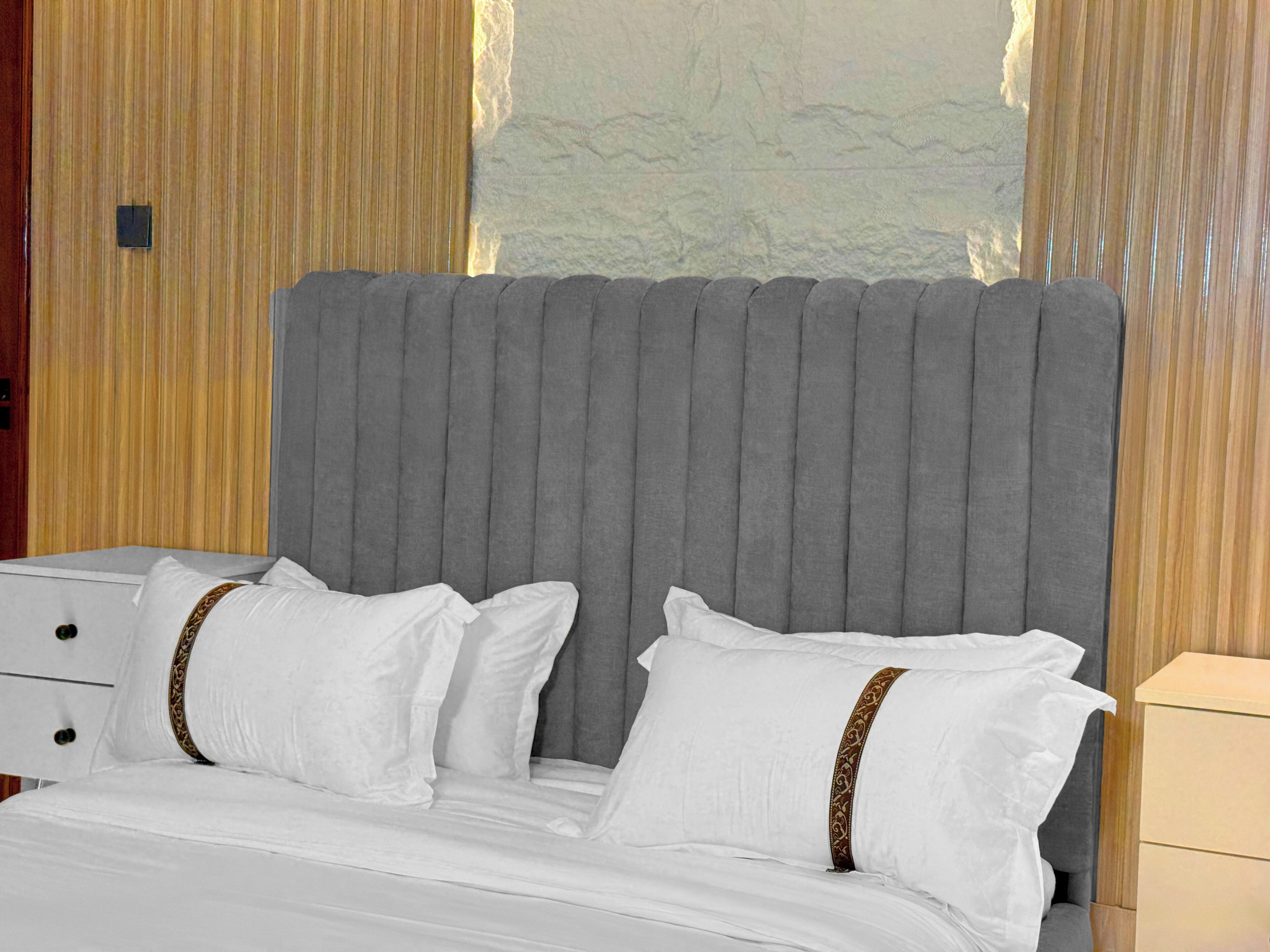 Un lit doté d’une tête de lit rembourrée de couleur grise, de draps blancs et d’oreillers décoratifs avec un bordure marron.