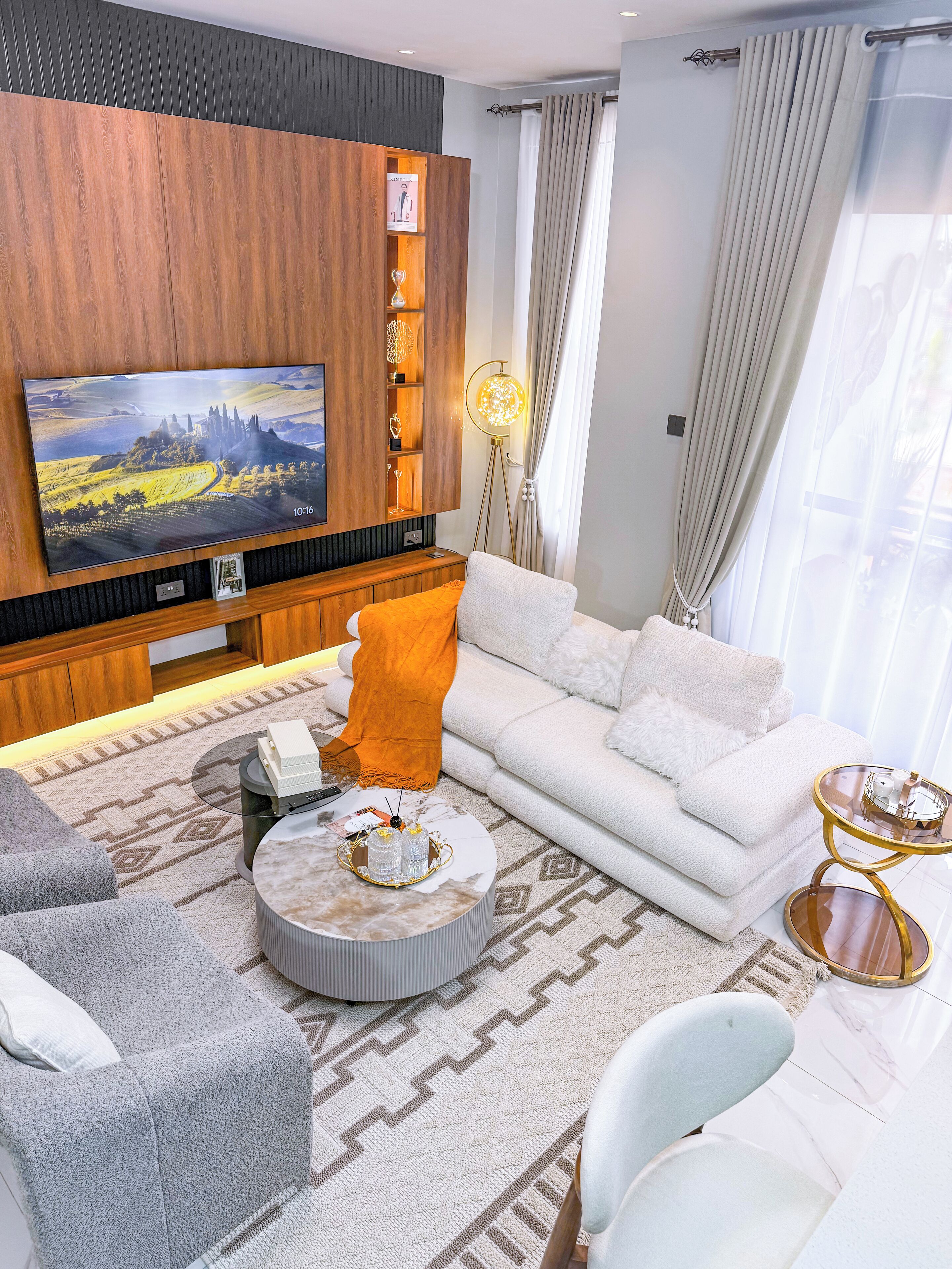 A modern living room with a large flat-screen TV mounted on a wooden wall, a white sofa, a grey armchair, a round coffee table with decorative items, and a floor lamp.