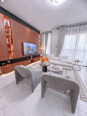 A modern living room with a flat-screen TV, a sofa, two grey poufs, a coffee table, and a decorative floor lamp. - Ivys Cozy Homes Kampala (Kampala)