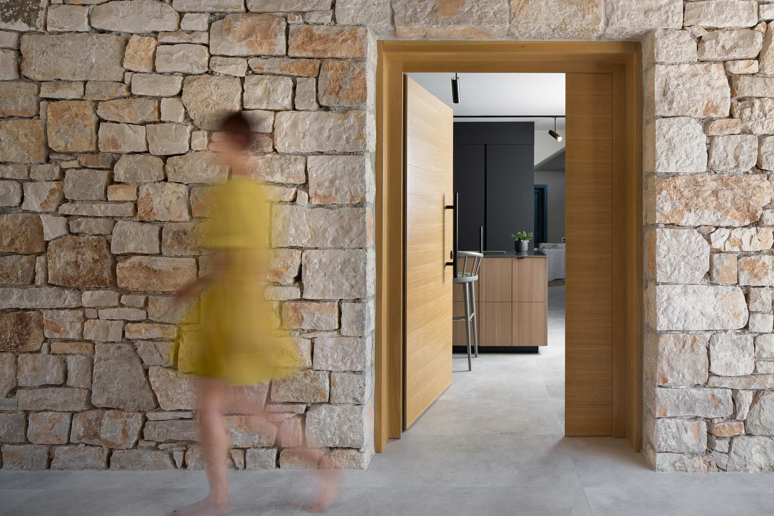 A room with a stone wall, a wooden door, and a view into another room with a kitchen area.