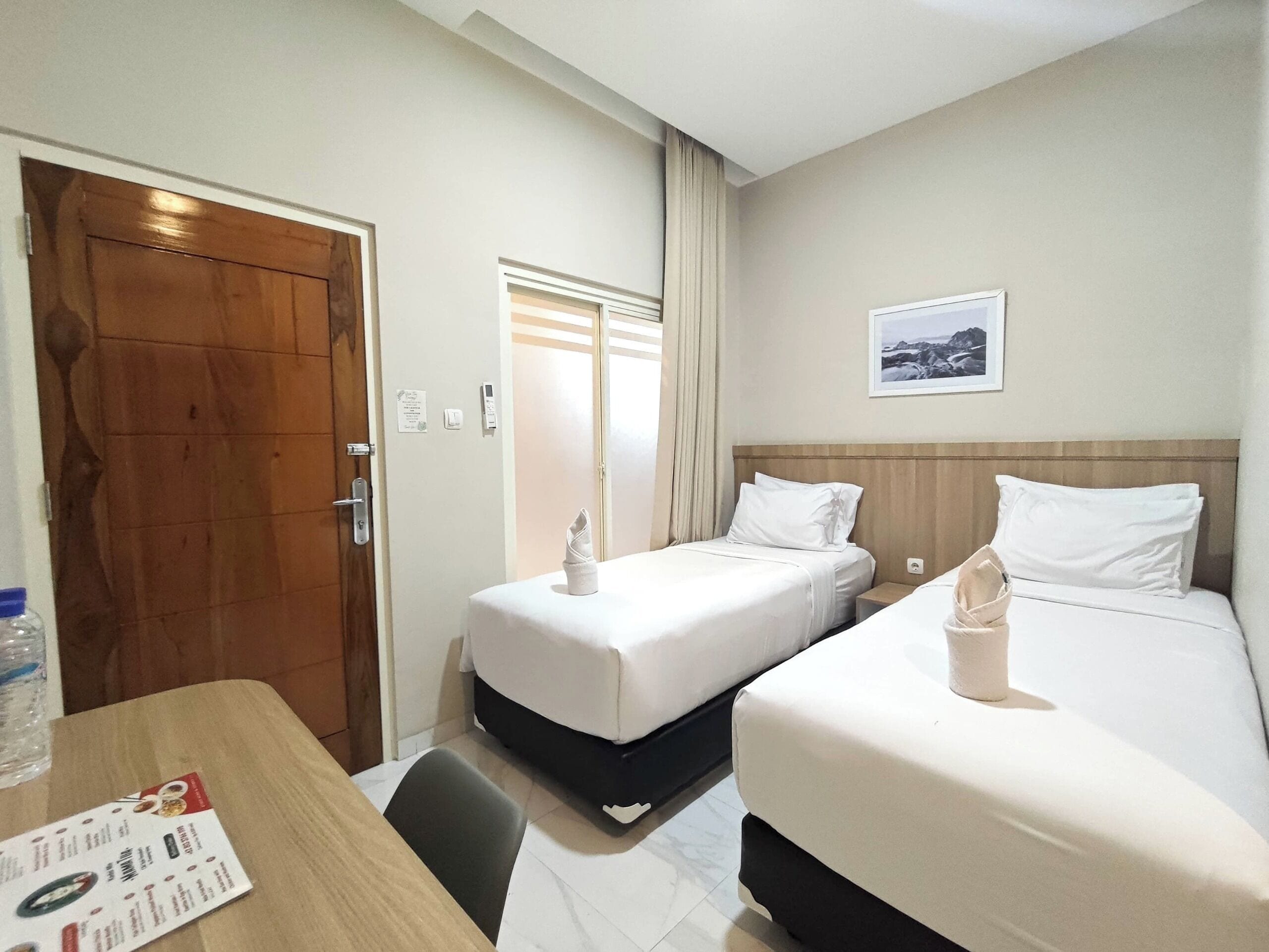 A hotel room with two single beds, a wooden door, a small table with a menu, and a framed picture on the wall.