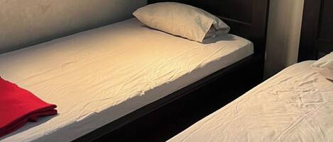 Una cama individual con cabecera de madera y almohada roja, una lámpara de pared y una ventana con contraventanas de madera.