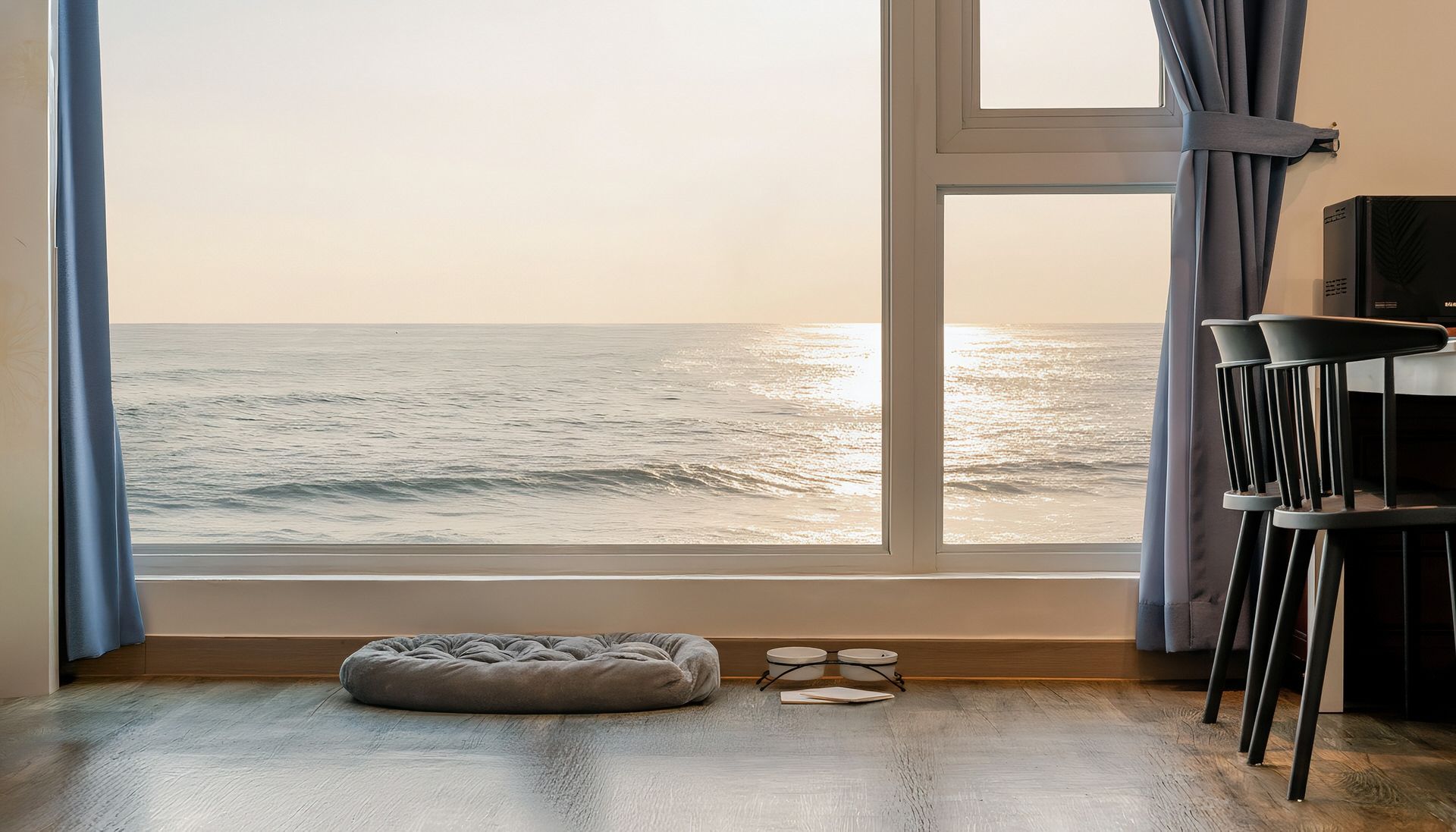 A room with a large window overlooking the sea, a grey bean bag chair, and a small table with a pair of glasses on it.