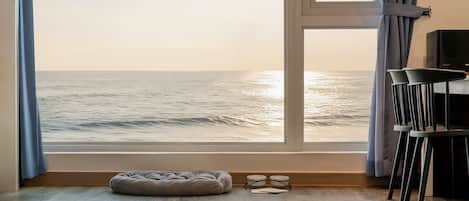 A room with a large window overlooking the sea, a grey bean bag chair, and a small table with a pair of glasses on it.