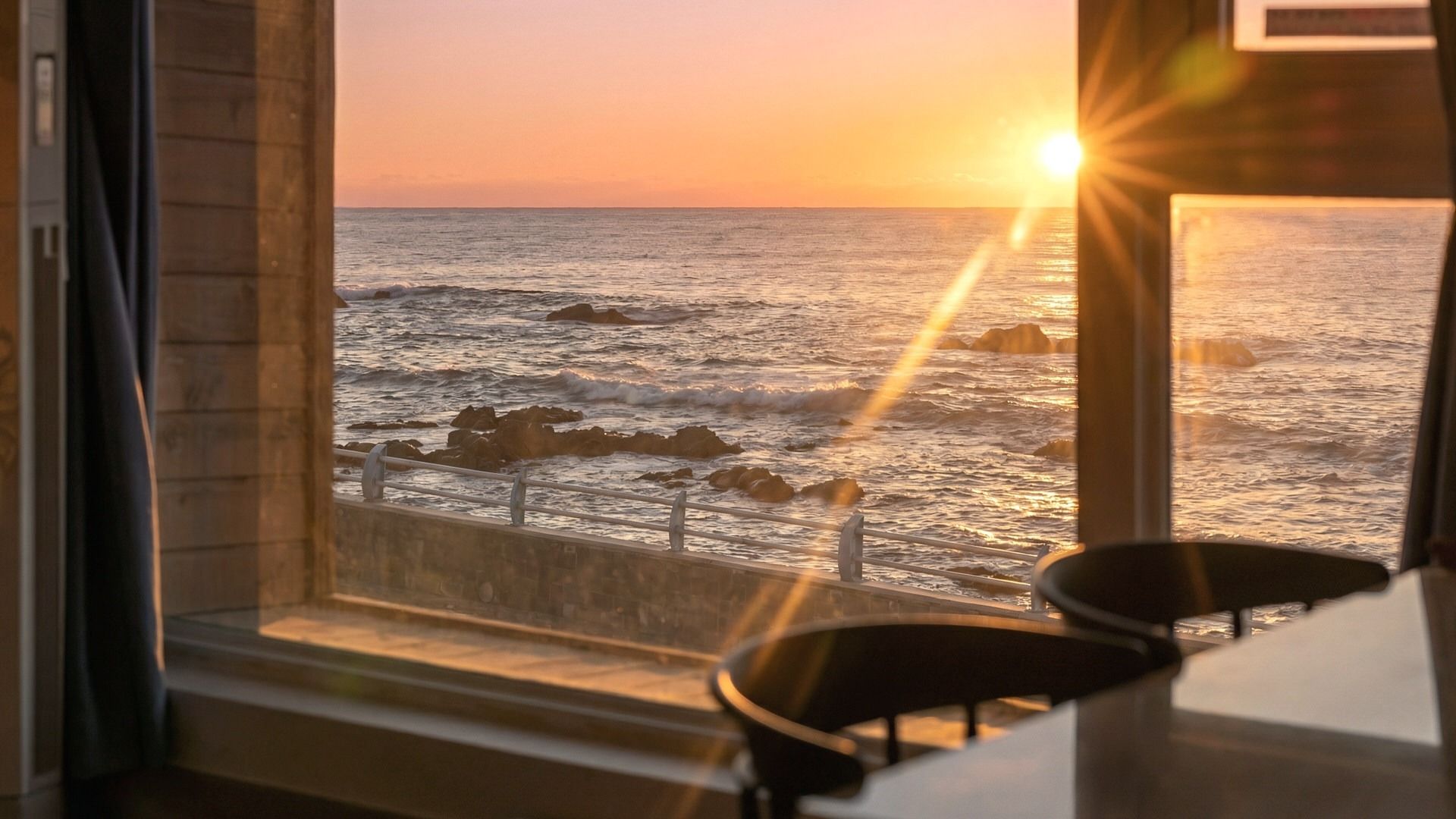 Vista del océano a través de una ventana con barandilla, una silla y una mesa.