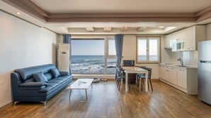 A modern living room with a sofa, coffee table, and dining area, a kitchen with a refrigerator, and a large window overlooking the sea. - Haru Stay Donghae (Donghae)