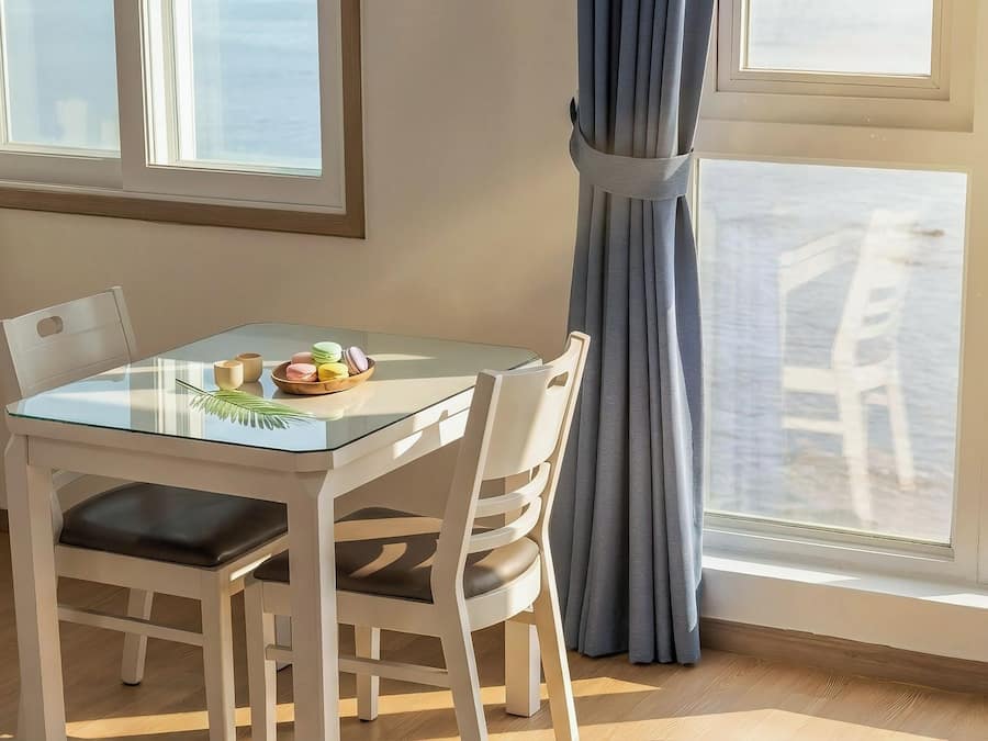 Una habitación con una mesa de vidrio, sillas blancas, vista a la playa y una ventana con cortinas.