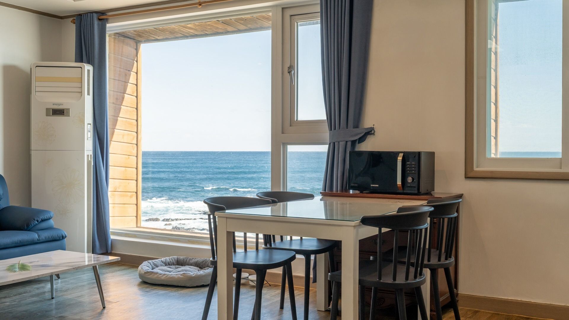 Una habitación con vistas al mar, una mesa de comedor con sillas, un sofá azul, un microondas y una cama pequeña para mascotas.