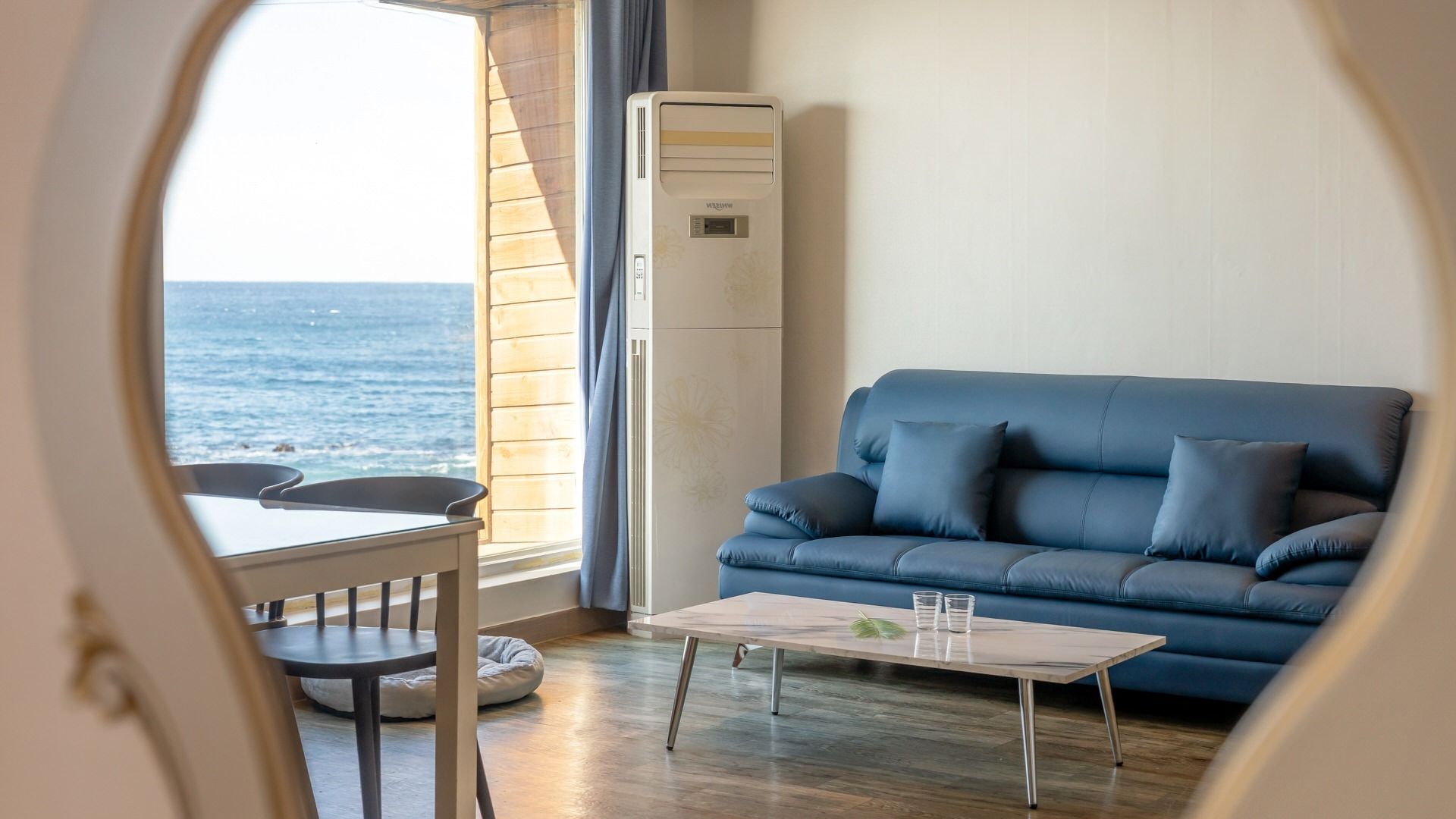 Ein Zimmer mit einem blauen Sofa, einem kleinen Tisch, einem Stuhl und Meerblick durch ein Fenster.