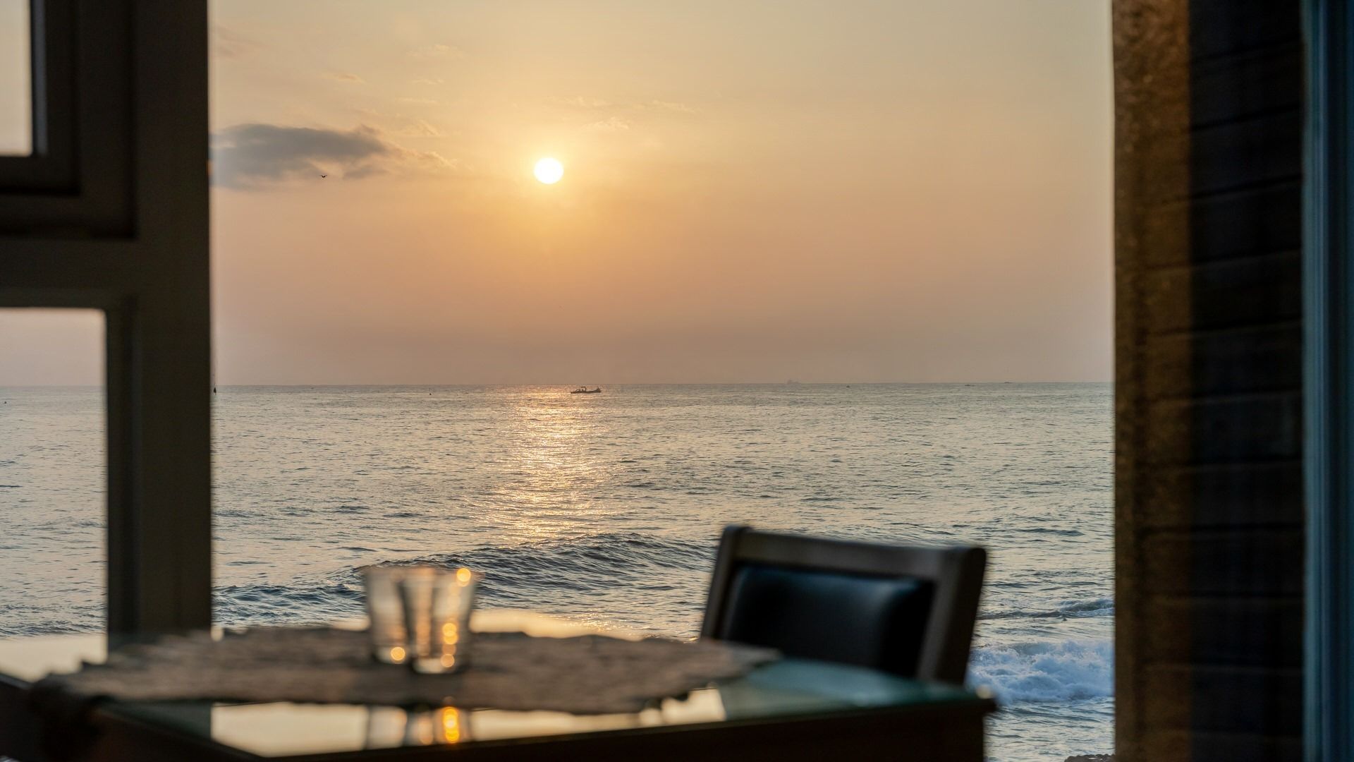 A table with a glass and chair, overlooking a calm sea with a boat and a setting sun.