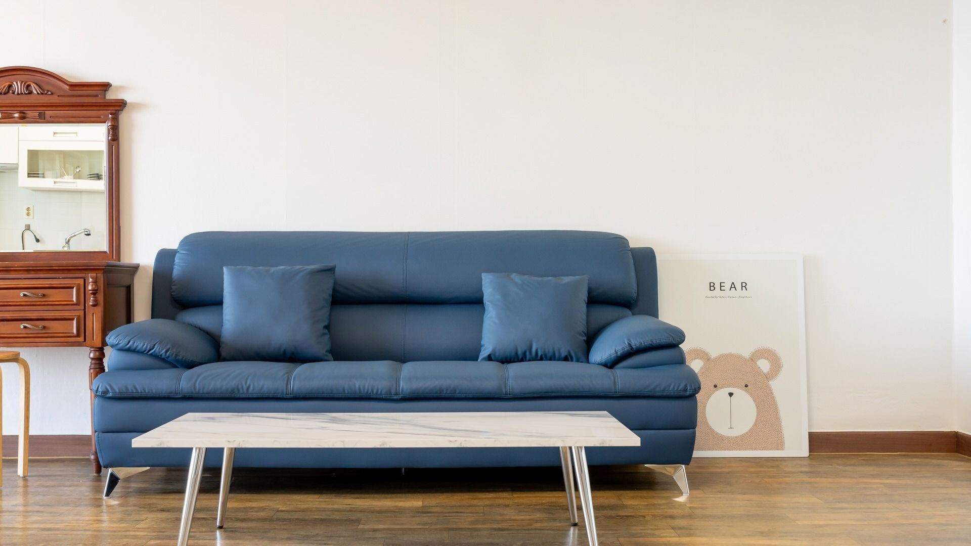 A blue sofa with two cushions, a wooden dresser, a marble coffee table, and a wall with a framed artwork.