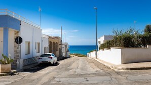 Una stretta via con un'auto bianca parcheggiata a sinistra, che conduce a un edificio con facciata bianca e un cielo azzurro sullo sfondo.