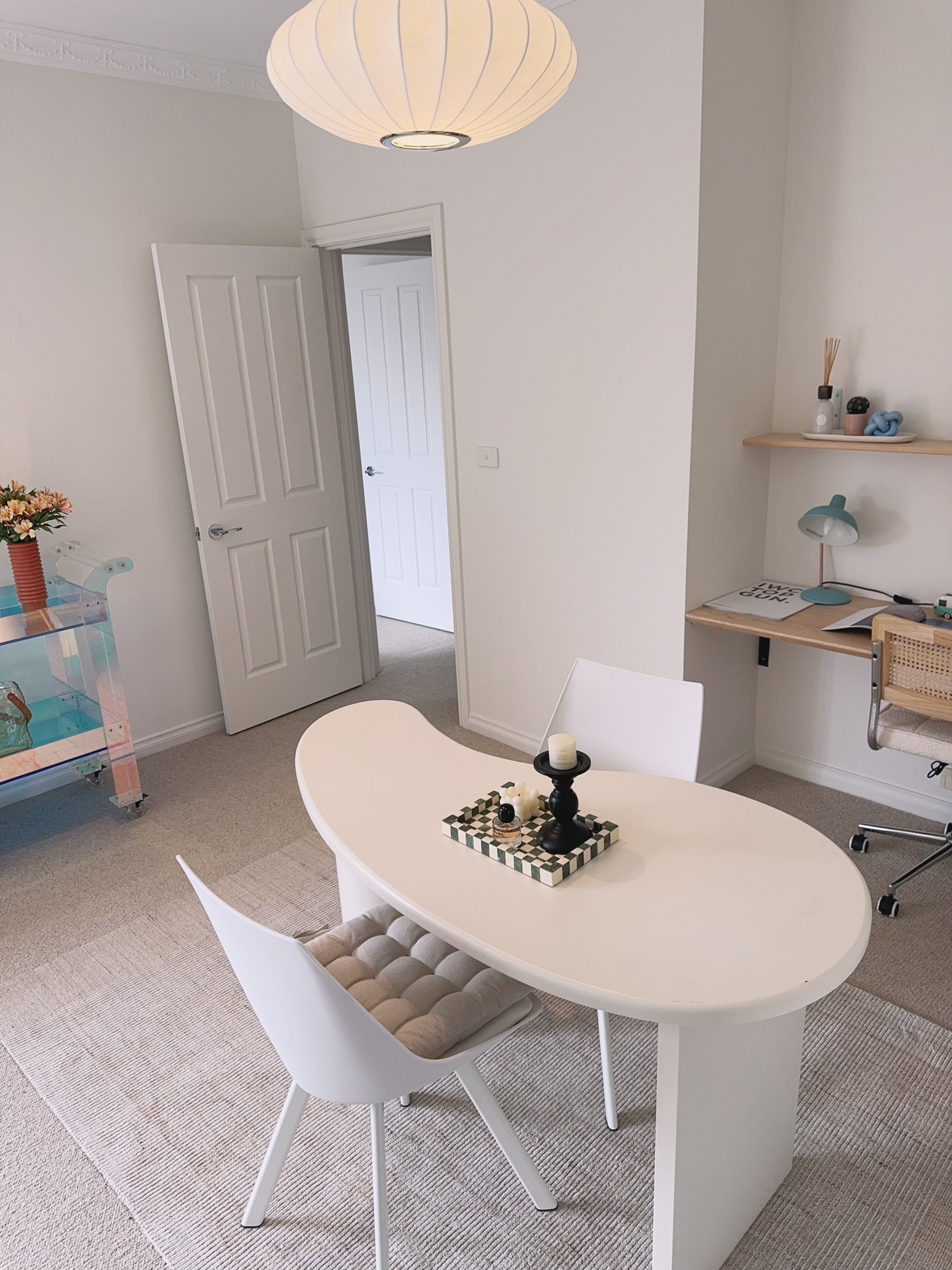 A modern, well-lit room with a white table, chairs, and a decorative lamp. A door leads to another room, and there is a shelf with small items.