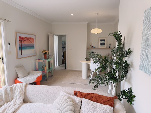 A modern living room with a white sofa, a coffee table, and a dining area with a white table and chairs. There is a plant in the corner and a painting on the wall.
