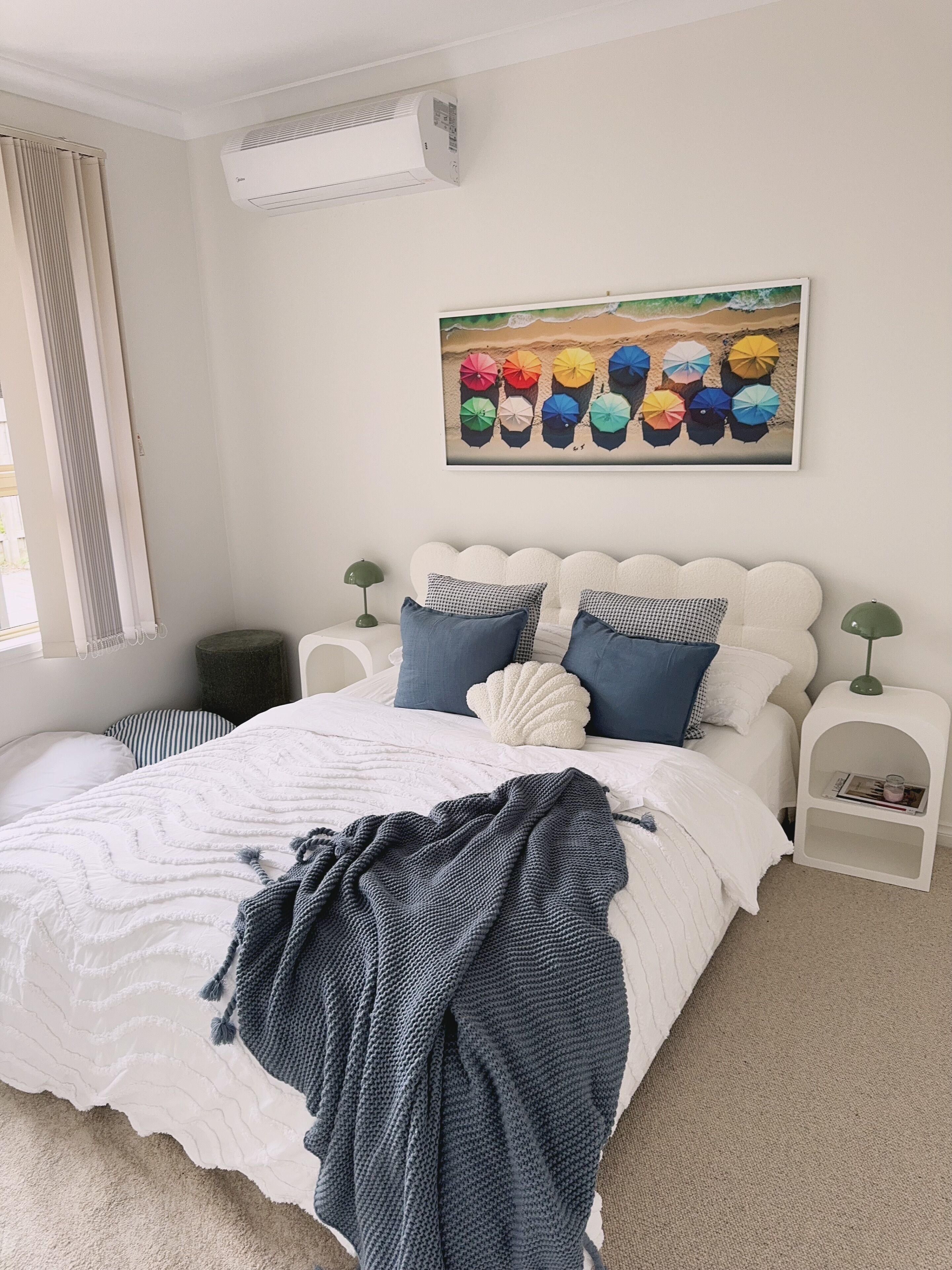 Een net opgemaakt bed met een wit- en blauw geborreld beddekleed, een blauwe gebreide plaid en een wandairconditioner.