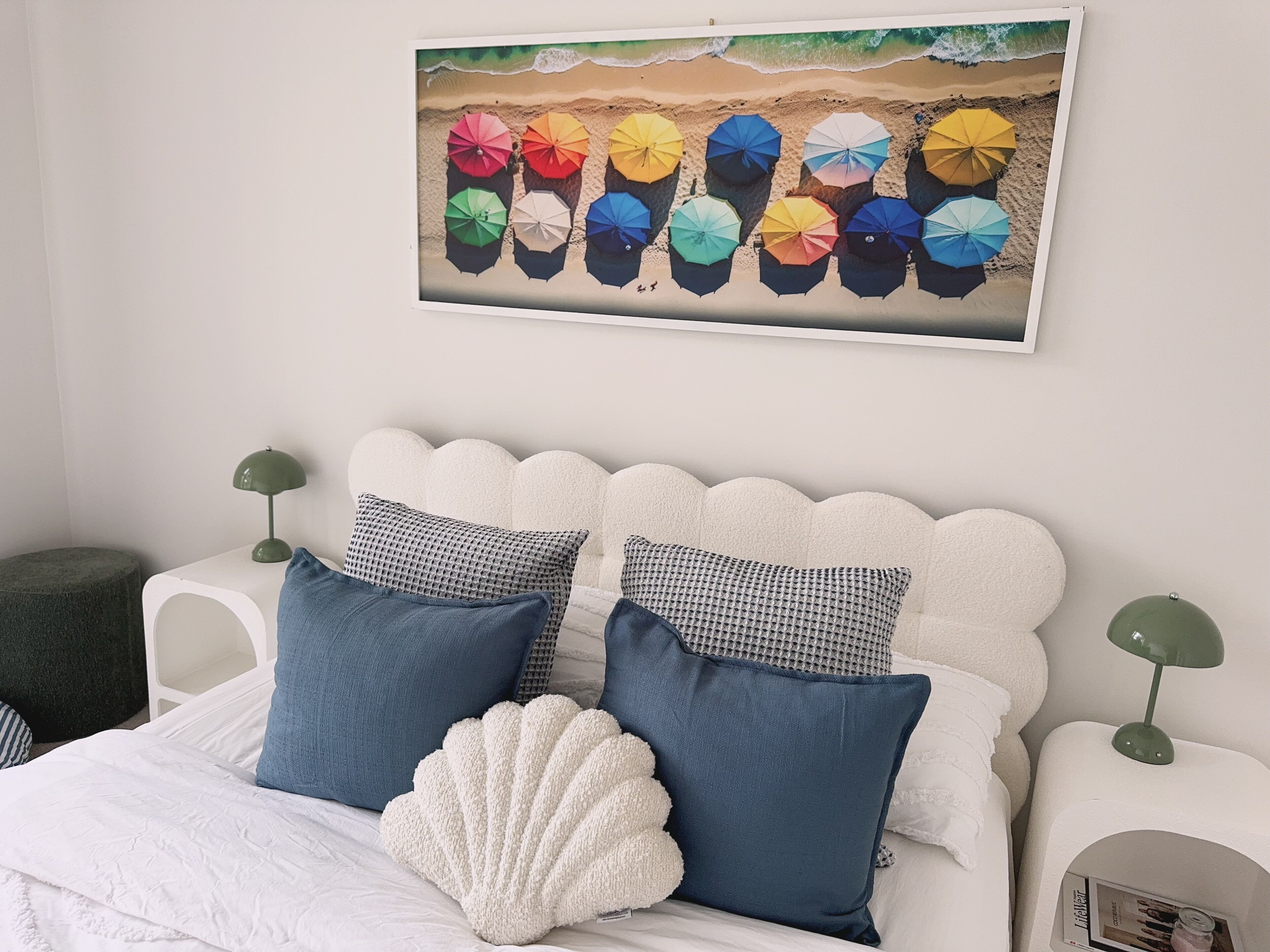 A neatly arranged bedroom with a white bed, two pillows, a white decorative shell, a wall-mounted painting of beach umbrellas, and two green bedside lamps.
