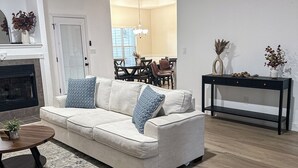 Ein Wohnzimmer mit einem beigefarbenen Ecksofa, einem Couchtisch, einem Teppich und einem Essbereich im Hintergrund.