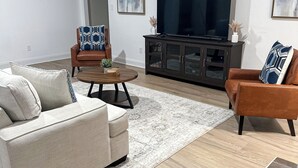 Ein Wohnzimmer mit einem beigen Sofa, zwei Sesseln, einem Couchtisch und einem Fernseher auf einem Schrank.