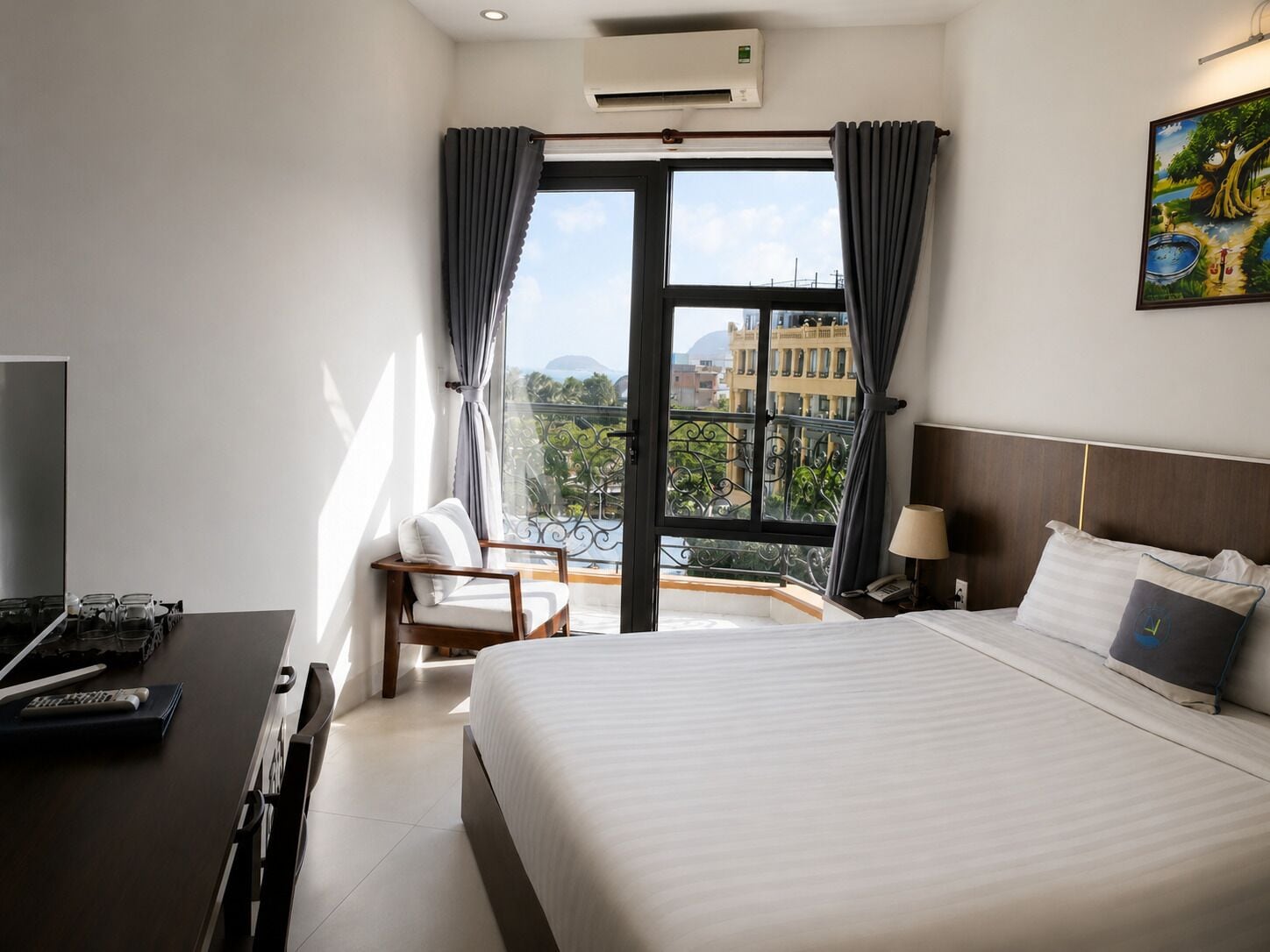 Una habitación de hotel con una cama, un escritorio con computadora, una silla, un ventanal con cortinas y un cuadro en la pared.