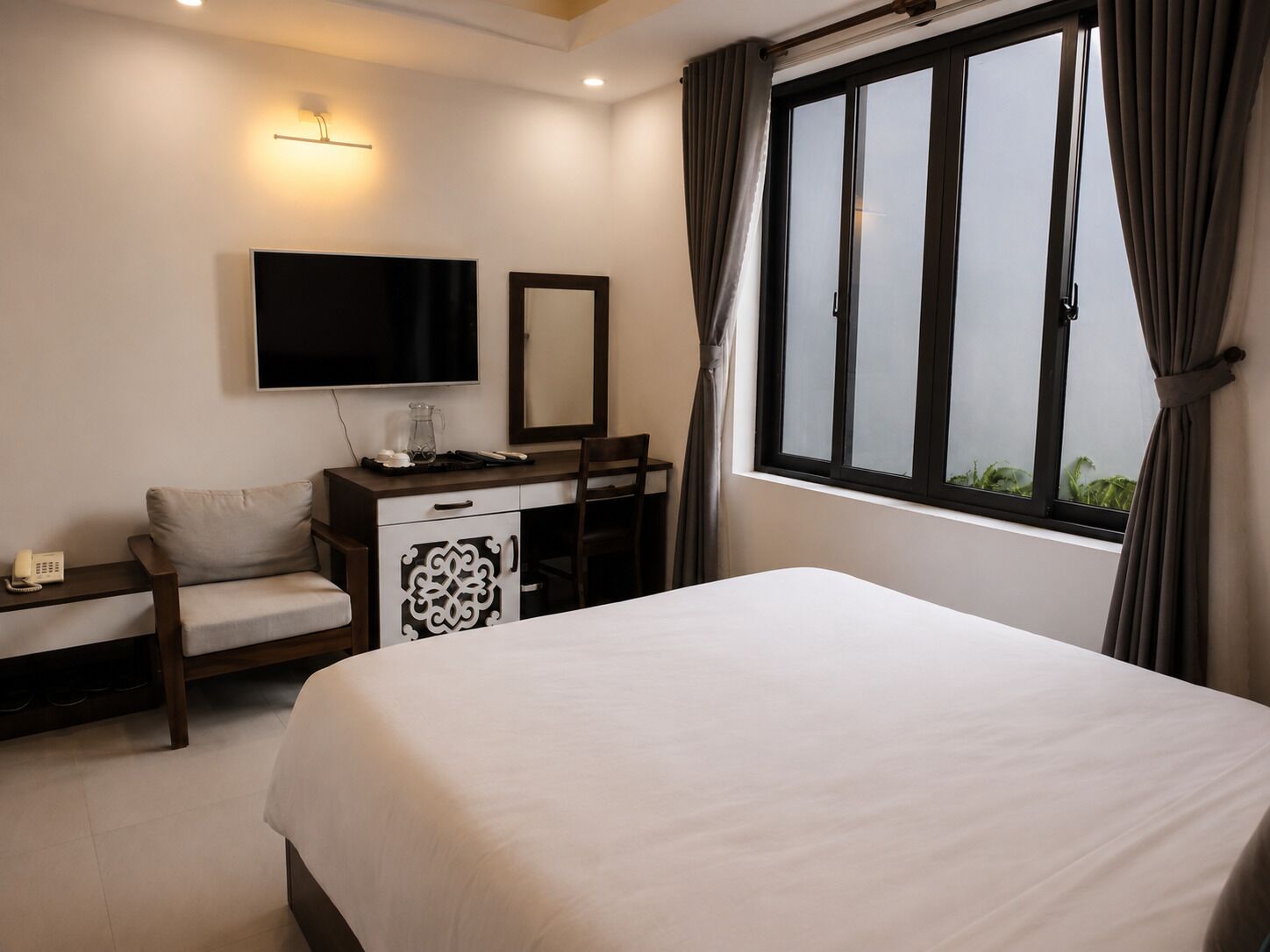Habitación de hotel con cama, escritorio con televisor de pantalla plana, silla, mesita de noche con lámpara y ventana con cortinas.