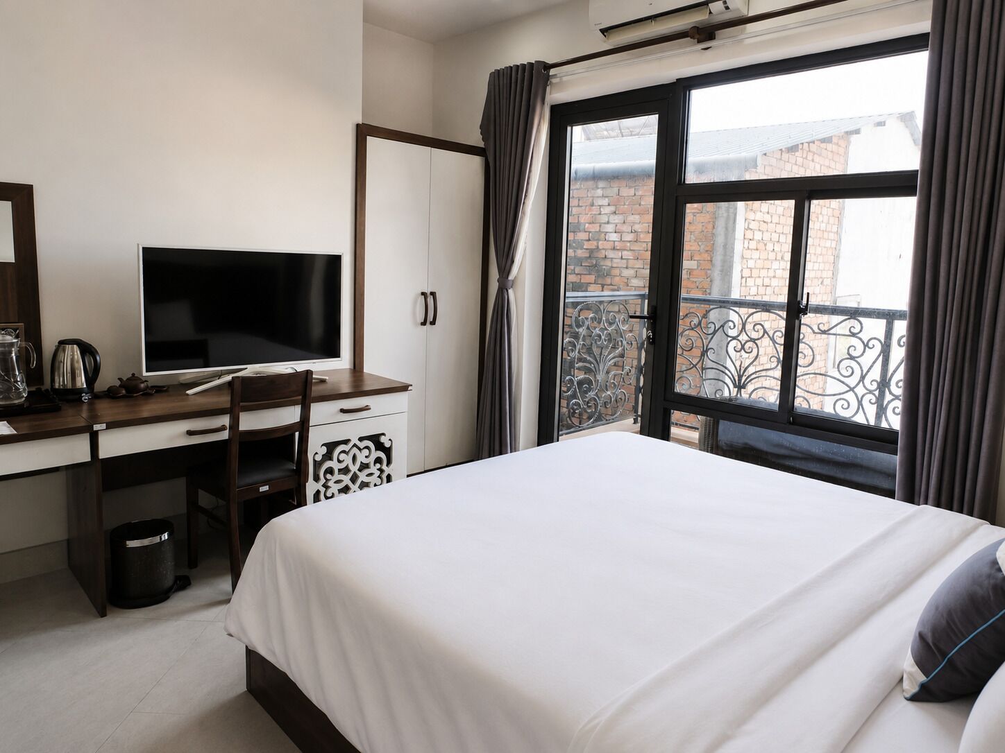 Un dormitorio con cama, escritorio con computadora, silla, ventana con cortinas y vista a un edificio de ladrillo.