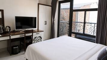 Un dormitorio con cama, escritorio con computadora, silla, ventana con cortinas y vista a un edificio de ladrillo.